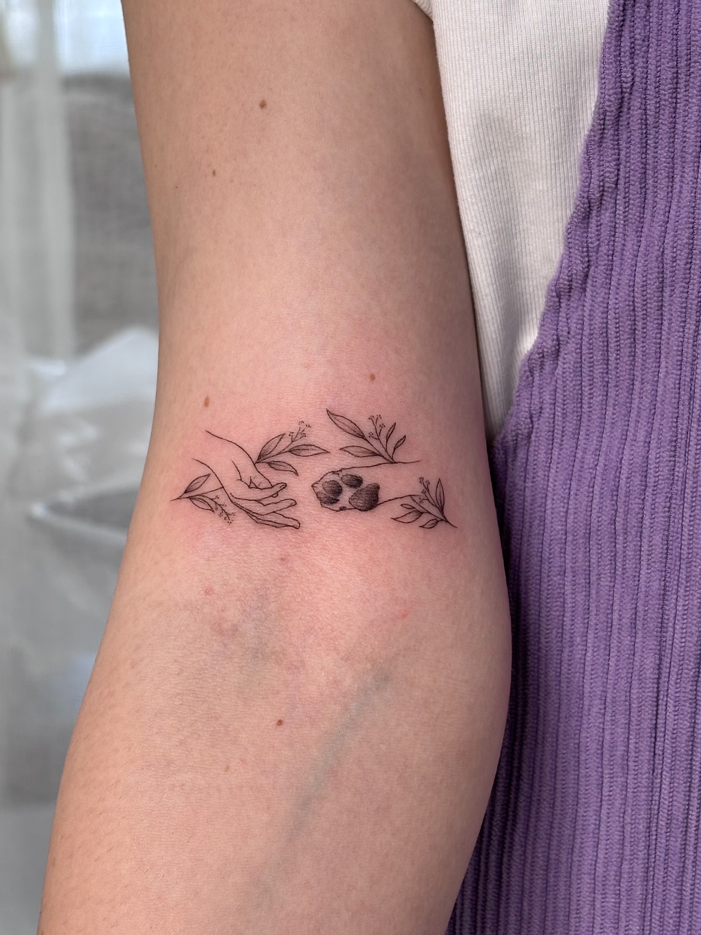 Fine line tattoo on an inner arm — a hand reaching toward a dog's paw print, framed by delicate leaves — by On Point Fine Line Tattoo Studio, Woodridge IL